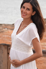 Bella mujer latina embarazada tocando barriga con vestido blanco al aire libre.