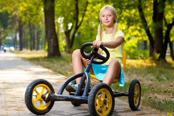 Fototapeta premium Beautiful smiling little girl riding toy car in summer city park