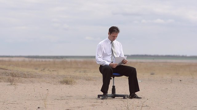 The man sits against the blue sky on a chair in the desert. The man watches documents in the black folder and thumbs through pages
