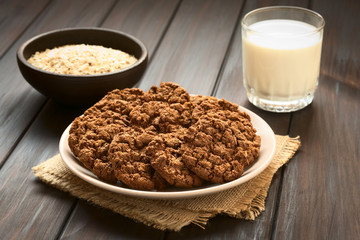 Chocolate oatmeal cookies on plate with a glass of milk and a bowl of oatmeal in the back, photographed with natural light (Selective Focus, Focus on the first cookies)