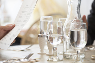 Close up of wine and water glasses and place settings at a table in a restaurant. A person's hand holding the menu.