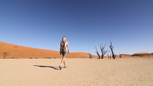 A girl walks across the salt pan of the Sossusvlei desert in Namibia, moving towards dead trees, with orange sand dunes and a blue sky in the background.