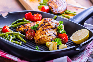 Grilled chicken breast in different variations with cherry tomatoes, green French beans, garlic, herbs, cut lemon on a wooden board or teflon pan. Traditional cuisine. Grill kitchen. 