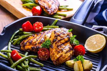 Grilled chicken breast in different variations with cherry tomatoes, green French beans, garlic, herbs, cut lemon on a wooden board or teflon pan. Traditional cuisine. Grill kitchen. 