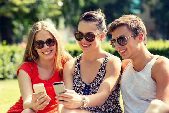 smiling friends with smartphones sitting in park