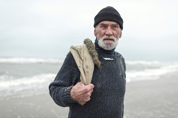 Senior beachcomber with burlap sack and woolen cap near sea.