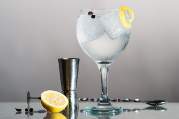 Gin Tonic with lemon and botanics in a balloon glass on grey background. With bar spoon and measure cup.