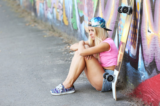 Young woman with skating board sitting on asphalt on painted wall background