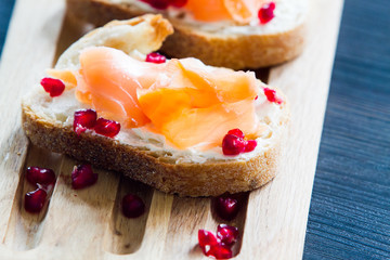 Salmon bruschetta with white cheese and pomegranates on wooden kitchen board over dark wooden background