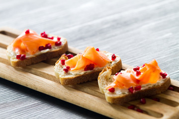 Salmon bruschetta with white cheese and pomegranates on wooden kitchen board over dark wooden background