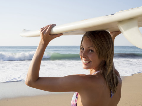 Rear view of smiling blonde sexy surfer young woman in bikini with white surfboard, wave on background