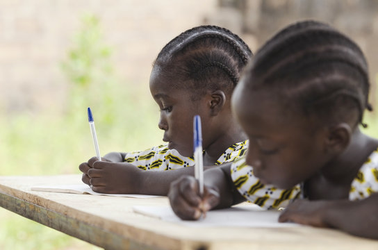 Schoolgirls repeating their lesson sitting in their desk at school in Bamako, Mali. Beautiful educational symbol. One girl's writing, the other one's slightly smiling.