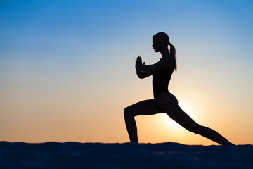 Silhouette young woman practicing yoga on sand at sunset