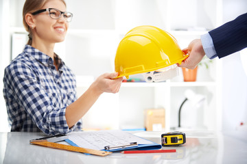 Smiling woman architect giving hardhat to male entrepreneur in office. Business agreement concept.