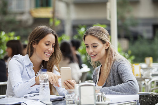 Two happy friends or sisters sharing a smart phone in a coffee shop terrace looking at device