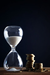 Hourglass and coins on wooden table on black background