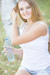 Selective focus on pretty young blond-haired woman wearing white T-shirt standing aside drinking water and smiling satisfied with her life. Park on background 