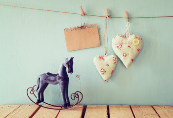 vintage rocking horse next to fabric hearts and empty card for adding text hanging on the rope on wooden floor. retro filtered image
