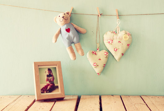 teddy bear over wood table next to photo frame with kid's old photography and fabric hearts. retro filtered image
