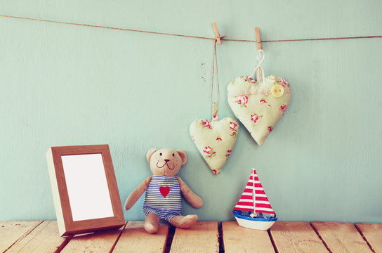 wooden boat toy and teddy bear over wood table next to blank photo frame and fabric hearts. retro filtered image
