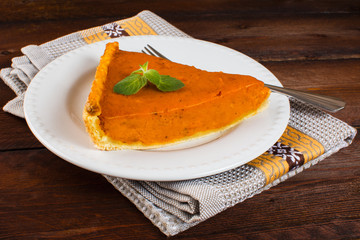 Thanksgiving pumpkin pie slice with mint on white plate, fork, linen napkin on dark wooden background