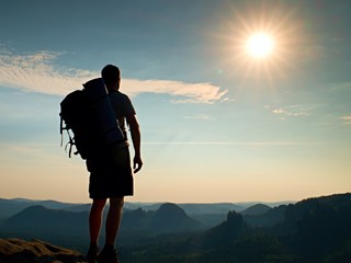 Tall tourist with poles in hand. Sunny evening in rocky mountains. Hiker with big backpack stand on...