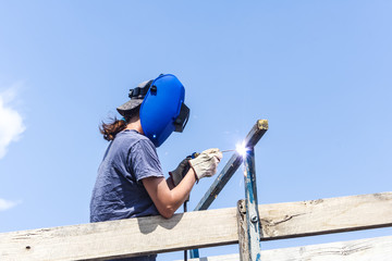 Female welder mending fence on farm wearing welding helmet showing flash spark. - obrazy, fototapety, plakaty