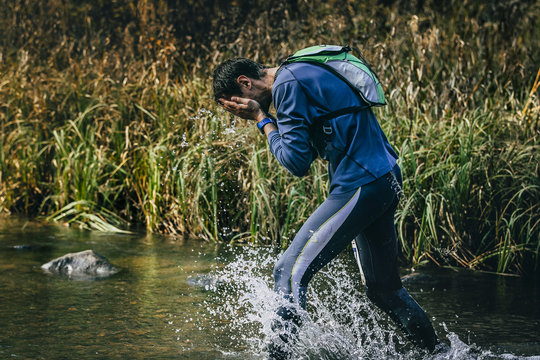 Male Athlete Running Across River. Rinses Face With Water