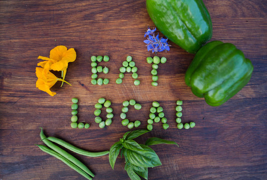 “Eat Local” phrase made of green peas on wooden background. Edible flowers, peppers, basil and green beans on a side. Organic vegetable produce at a farm.
