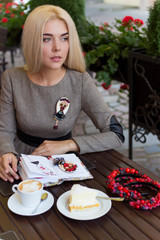 beautiful blonde girl sitting at cafe with Cup of coffee and cake works and draws sketches in a notebook