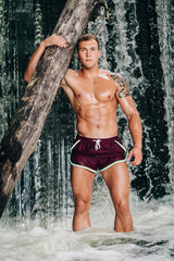 Shirtless muscled handsome young man with log on waterfall