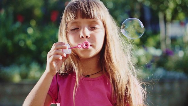 Cute little girl is blowing soap bubbles in the garden outdoors on a sunny day. Slow Motion 240 fps. Happy childhood concept. Child is playing in bright sun light. 