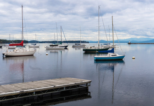 sail boats moored in lake  with breakwater and lighthouse seen before the Adirondack mountains in the distance 
