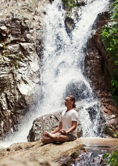 sporty young man practicing yoga near a waterfall in the mountai