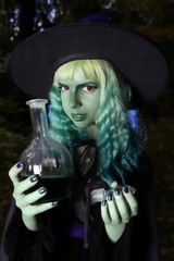 Young girl with green hair and flask with a potion in suit of witch in forest. Halloween time