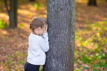 Little girl playing hide and seek near the tree in autumn park