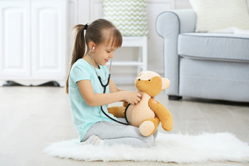 Little cute girl with stethoscope and teddy bear sitting on carpet, on home interior background