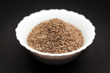 Organic Cumin seed (Cuminum cyminum) in white ceramic bowl on dark background.
