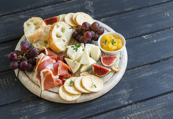 delicious appetizer to wine - ham, cheese, grapes, crackers, figs, nuts, jam, served on a light wooden board, and two glasses with white wine on  wooden surface