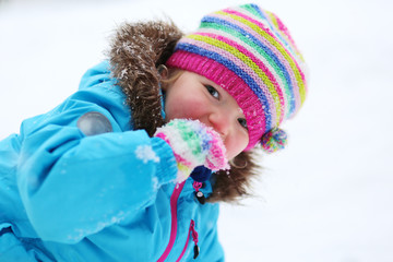 Portrait of beautiful toddler girl playing outdoors with snow. Happy little child wearing colorful knitted hat and blue coat enjoying winter day int he park.