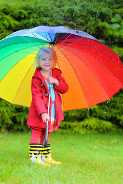 Little child with colorful umbrella playing in the garden during the rain. Cute toddler girl having fun outdoors by rainy fall weather. Preschooler in yellow wellies boots walking in park.
