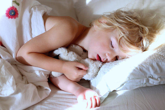 Sweet Little Child Sleeping In Bed. Healthy Kid, Blonde Toddler Girl, Resting In Bed In White Sunny Bedroom.