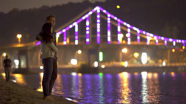 The couple (pair) stand and hug on the beach picturesque embankment by night city and bridge lights background. Shot with Red Cinema Camera