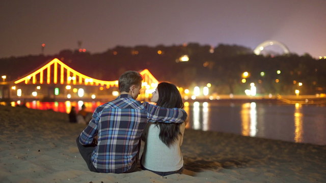The couple (pair) sit on the beach picturesque embankment by night city and bridge lights background. Shot with Red Cinema Camera
