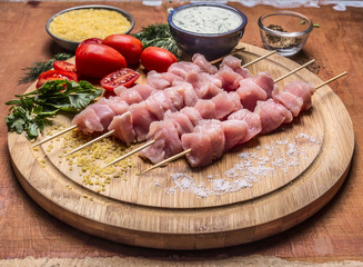 Raw chicken pieces on skewers on a chopping board with herbs vegetables and sauce on wooden background close up