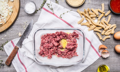 kitchen table preparation of raw minced meat chopped onion seasoning eggs tomato sauce penne kitchen knife On a white napkin on wooden background top view close up