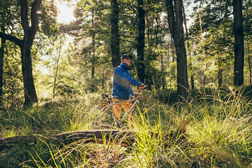Backlit retired man standing in forest with bicycle.