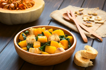 Pumpkin and chard salad with croutons served in wooden bowl, photographed on dark wood with natural light (Selective Focus, Focus in the middle of the salad)