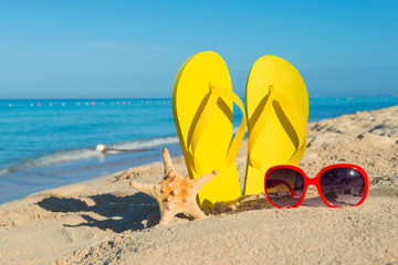 Fototapeta na wymiar Women's red sunglasses and yellow flip-flops on sandy seashore. Travel by sea. Beach vacation