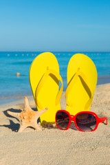 Fototapeta na wymiar Travel by sea. Beach vacation. Red sunglasses and yellow flip-flops on sandy seashore.